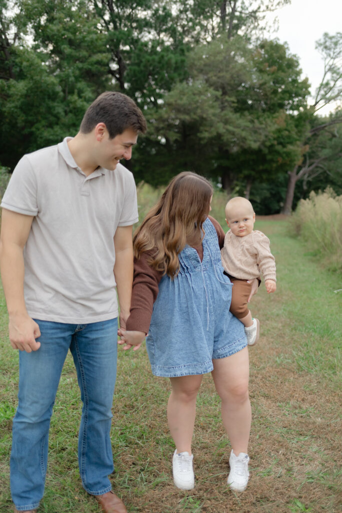 family session in a field, parents holding hands looking at baby 